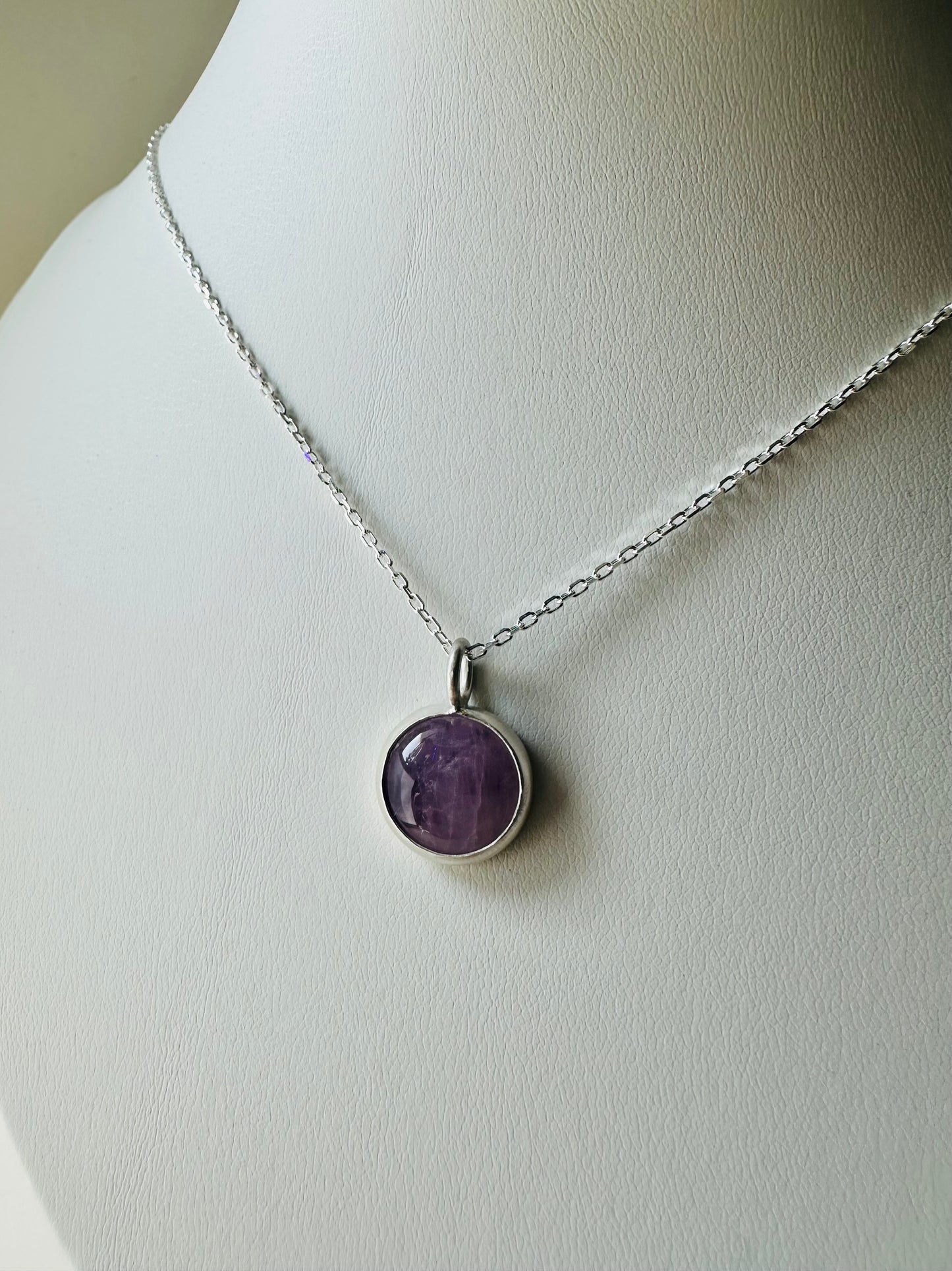 A silver chain necklace with a round bezel-set purple stone pendant, displayed on a white jewelry bust.
