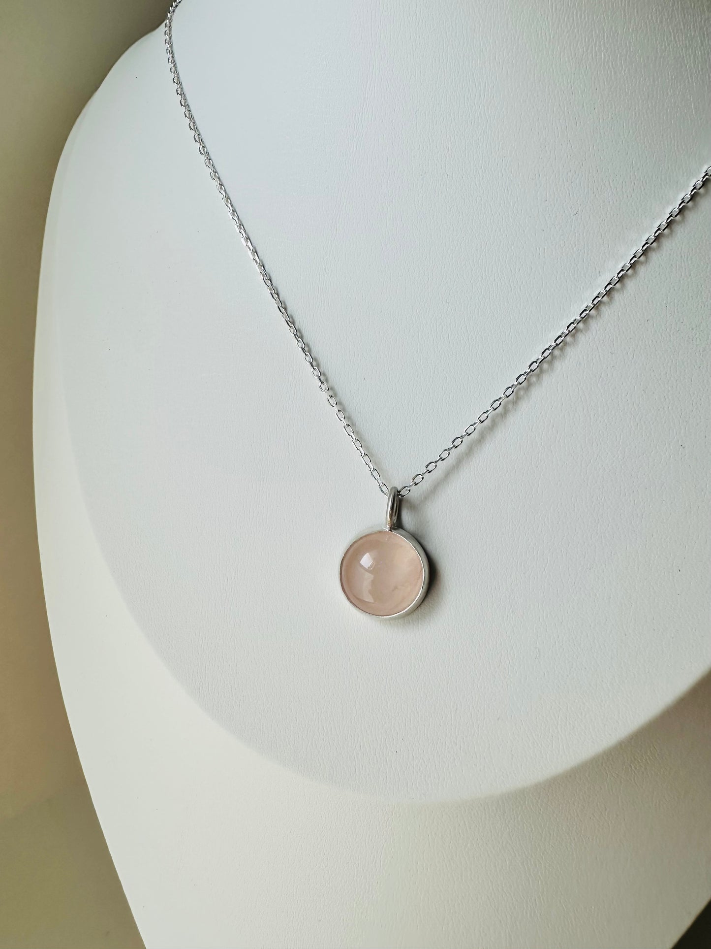 A silver chain necklace with a round, pale pink stone pendant displayed on a white jewelry bust.