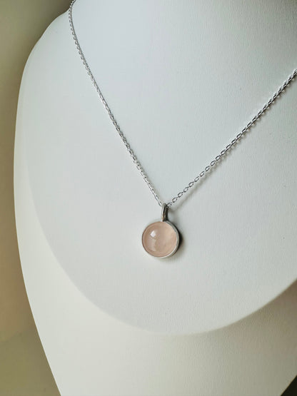A silver chain necklace with a round, pale pink stone pendant displayed on a white jewelry bust.