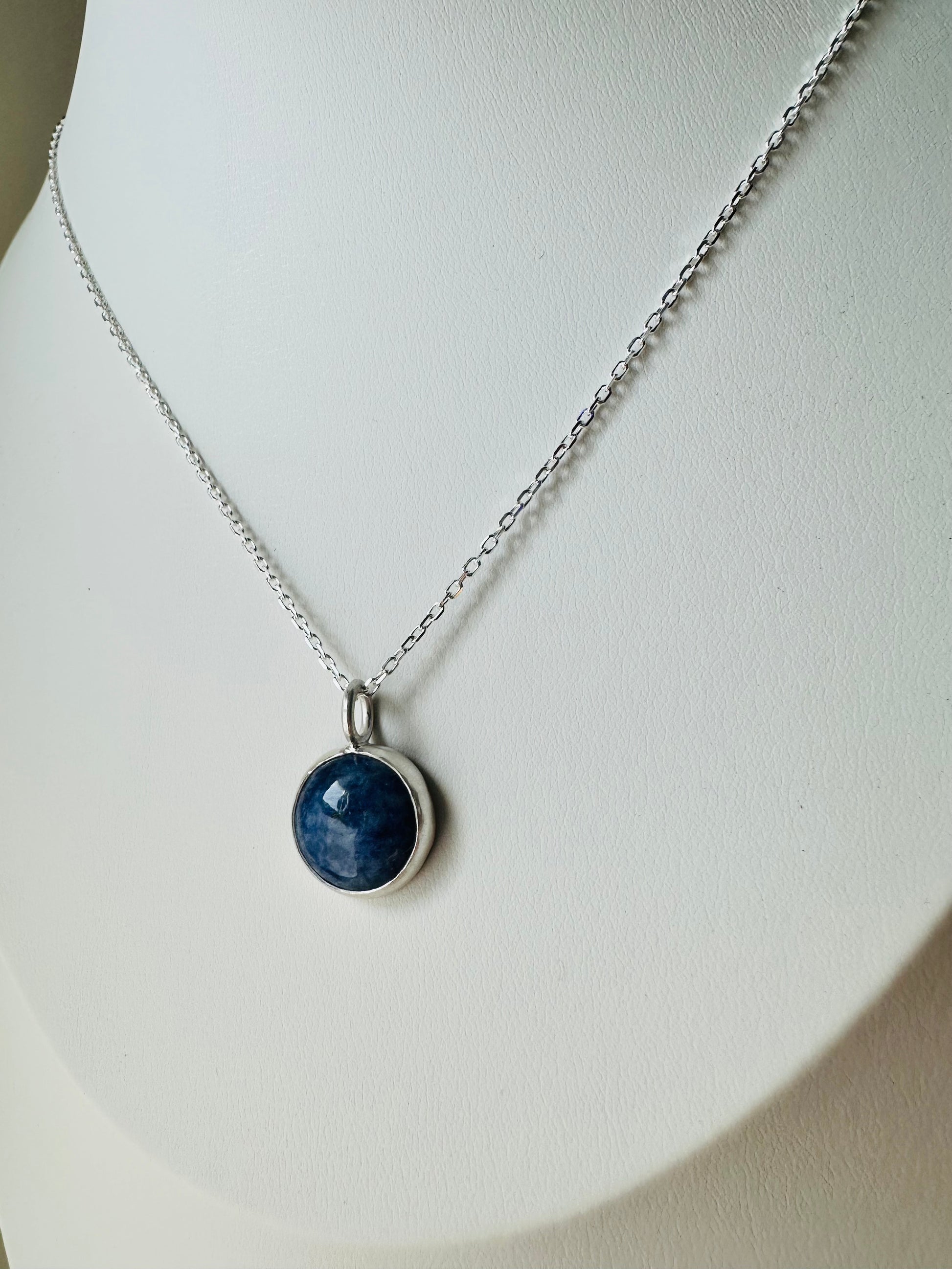 A silver chain necklace with a round dark blue stone pendant in a silver bezel setting, displayed on a white jewelry bust.