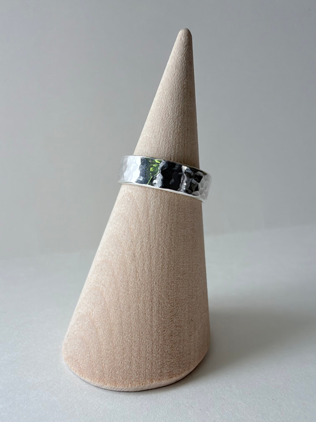 A hammered silver band ring displayed on a light wood cone jewelry stand against a neutral gray background.