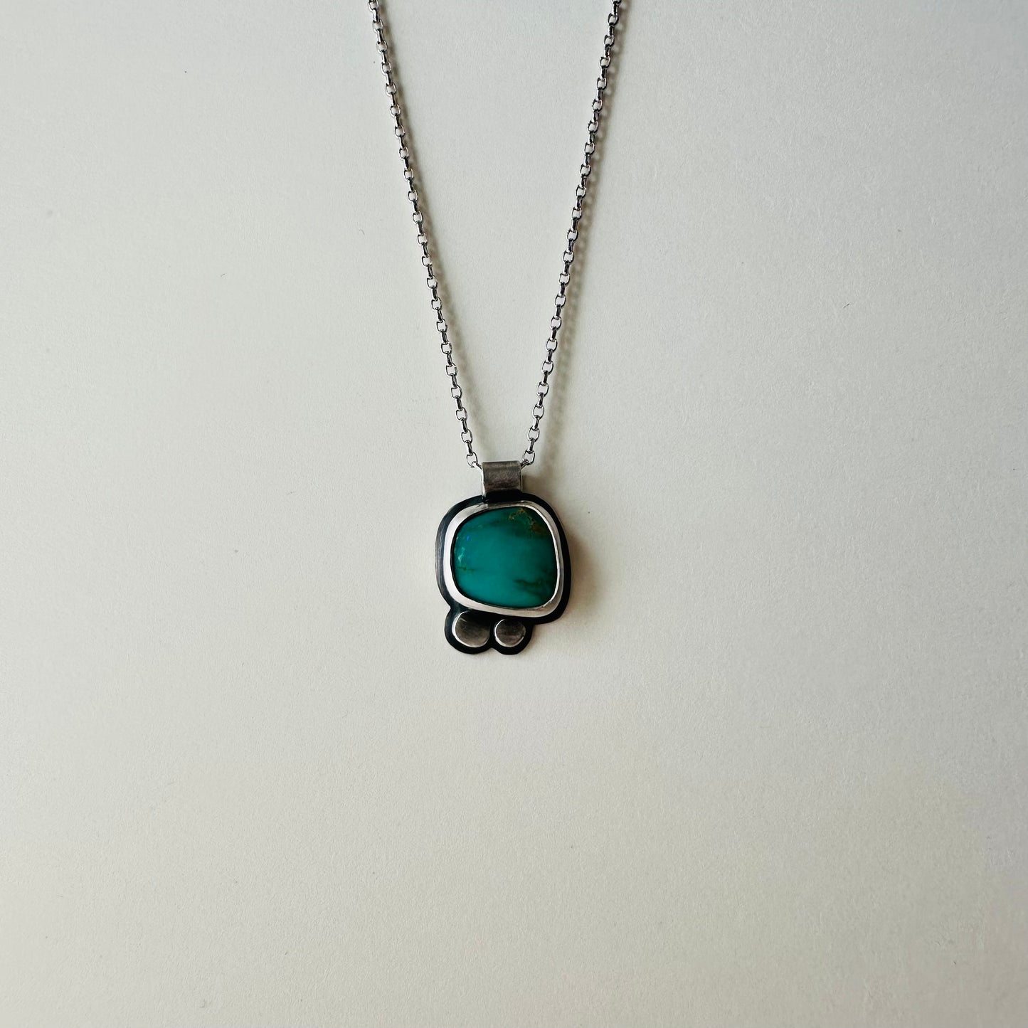 A silver chain necklace with a small pendant featuring a green stone in a dark metal setting, photographed against a plain light background.