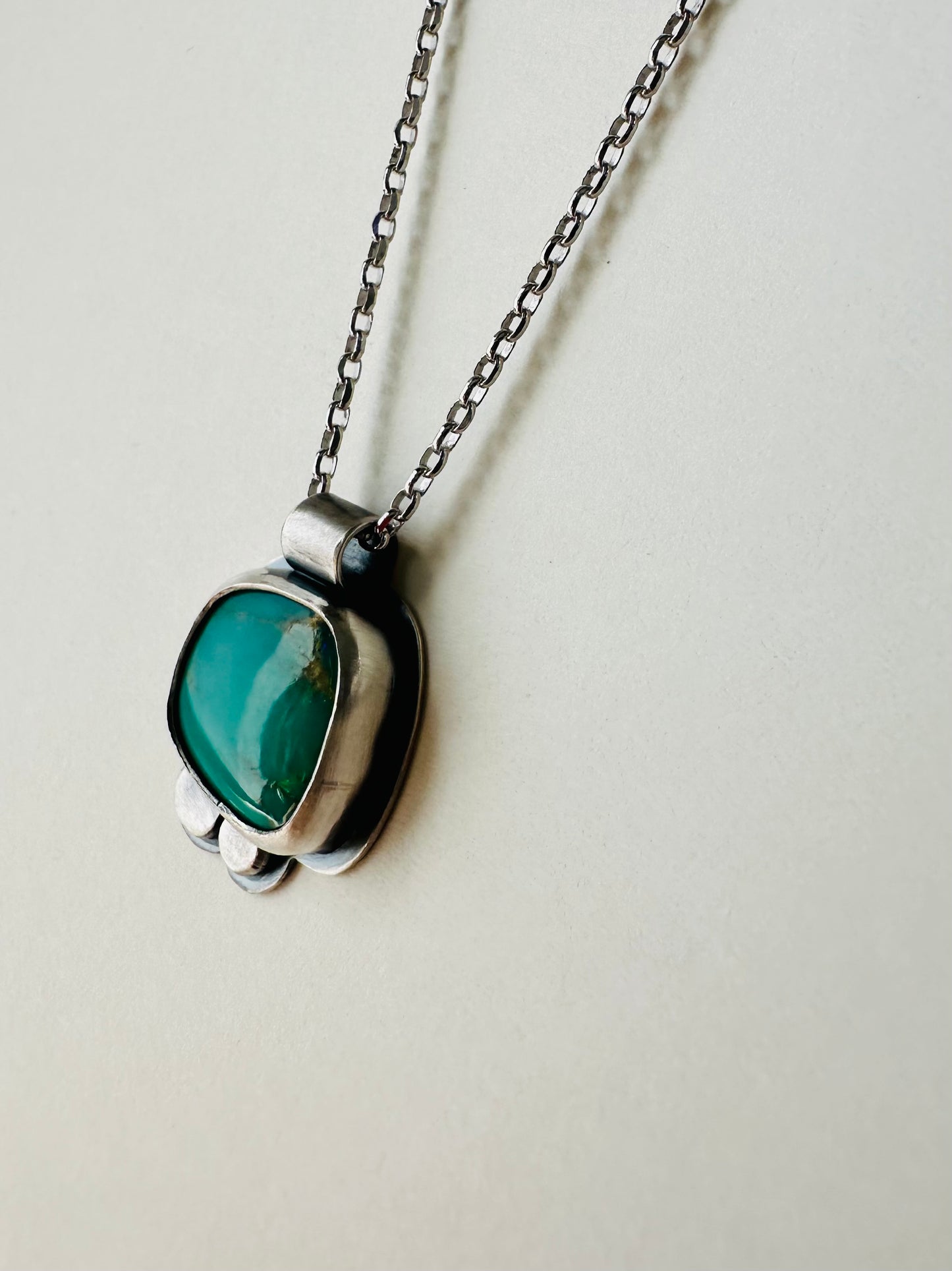 A silver pendant necklace featuring a rounded, organic-shaped green stone, hanging from a silver chain against a plain off-white background.