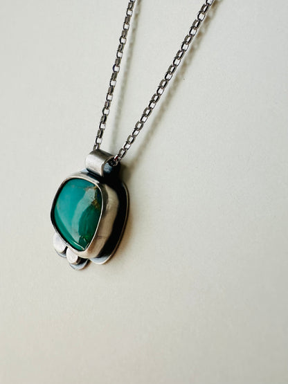 A silver pendant necklace featuring a rounded, organic-shaped green stone, hanging from a silver chain against a plain off-white background.