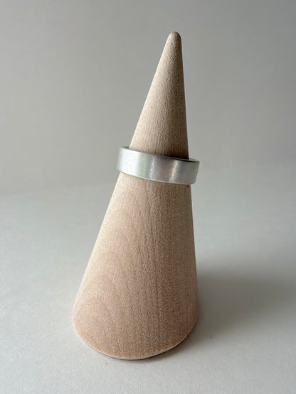 A wide, matte silver band ring displayed on a light wood cone jewelry stand against a neutral gray background.
