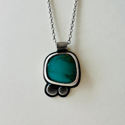 A silver pendant necklace featuring a vibrant turquoise colored stone in a rounded, handcrafted setting with two small silver circles below, hanging from a silver chain against a light off-white background.