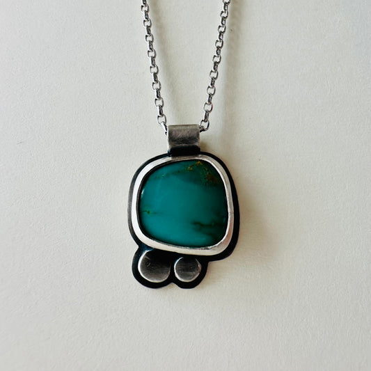 A silver pendant necklace featuring a vibrant turquoise colored stone in a rounded, handcrafted setting with two small silver circles below, hanging from a silver chain against a light off-white background.
