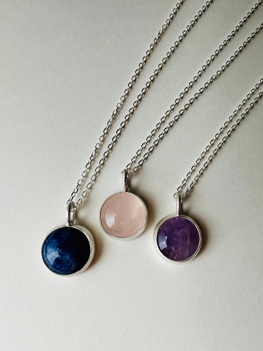 Three silver chain necklaces with round bezel-set gemstone pendants in navy blue, pale pink, and purple, arranged diagonally on a light cream background.