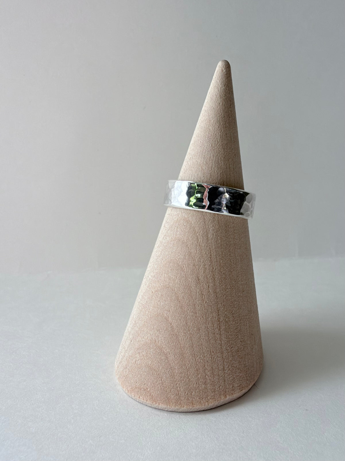 A polished thick silver hammered ring band displayed on a wooden cone stand against a soft gray background.