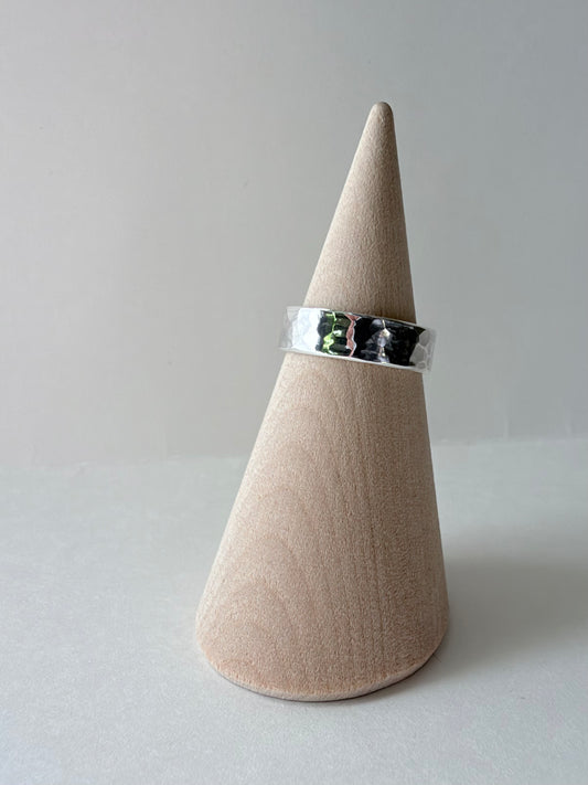 A polished thick silver hammered ring band displayed on a wooden cone stand against a soft gray background.