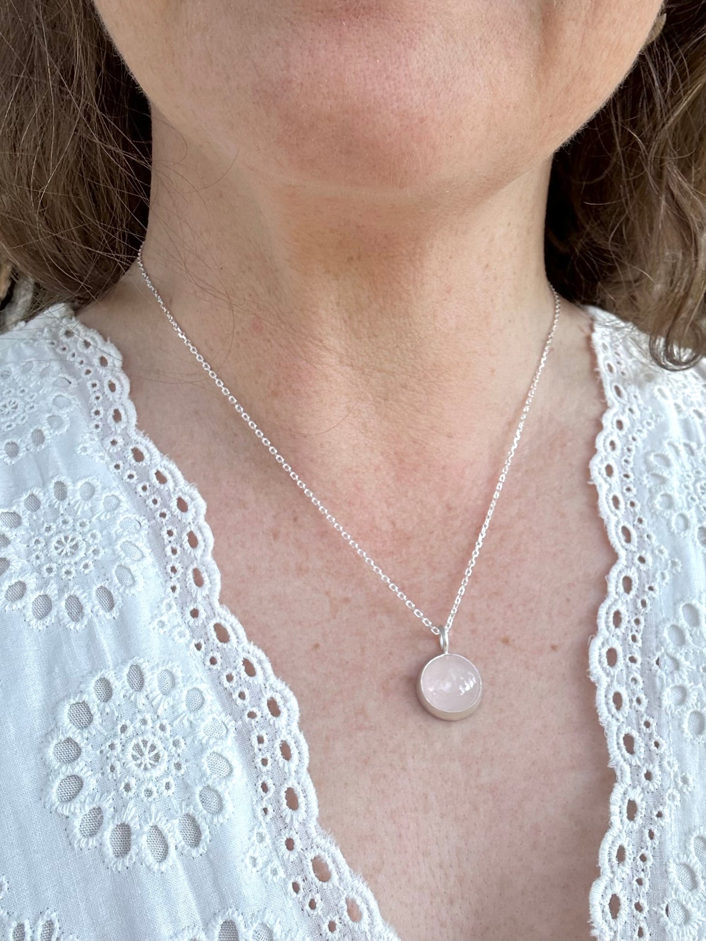 A close-up of a woman wearing a delicate silver chain necklace with a round, pale pink stone pendant, paired with a white lace-trimmed top.