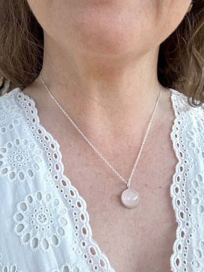 A close-up of a woman wearing a delicate silver chain necklace with a round, pale pink stone pendant, paired with a white lace-trimmed top.