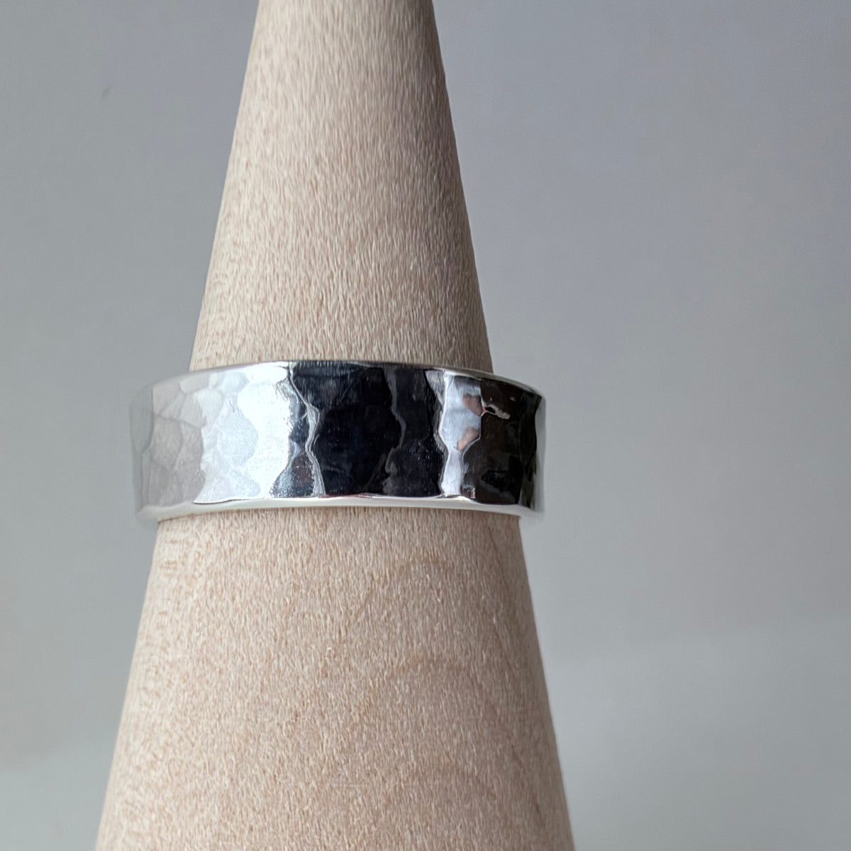 A hammered sterling silver band ring displayed on a light wood cone stand against a neutral gray background.