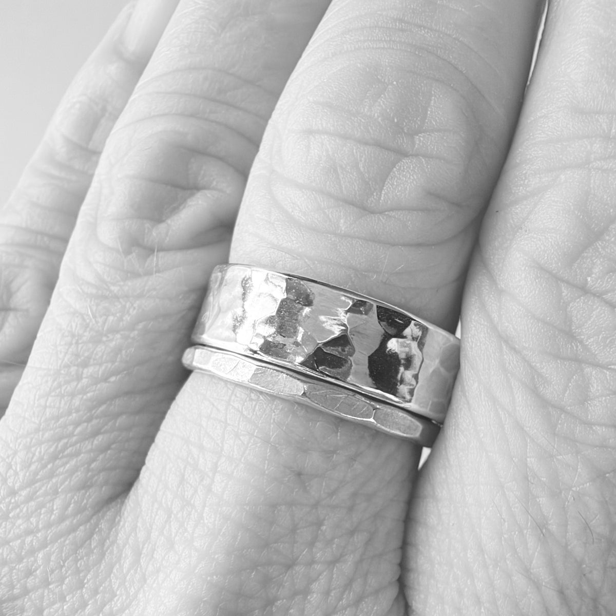A close-up of a silver hammered band ring worn on a finger, with a second textured silver band underneath, shown in black and white.