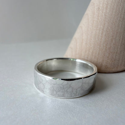 A silver hammered band ring resting on a light gray surface next to a wood ring cone.