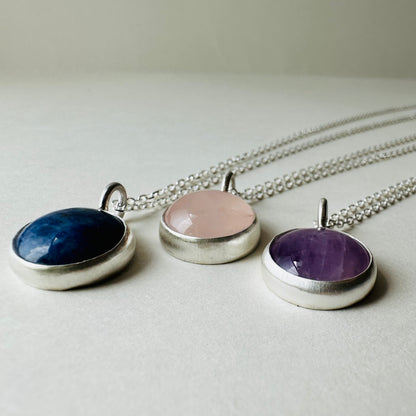 Three silver pendant necklaces with round cabochon gemstones in navy blue, pale pink, and purple, arranged on a light gray surface.