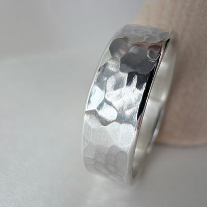 A hammered silver band ring resting on a light-colored textured surface against a white background.