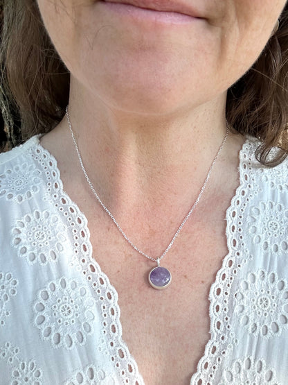 A close-up of a woman wearing a delicate silver chain necklace with a round purple gemstone pendant, paired with a white lace-trim top.
