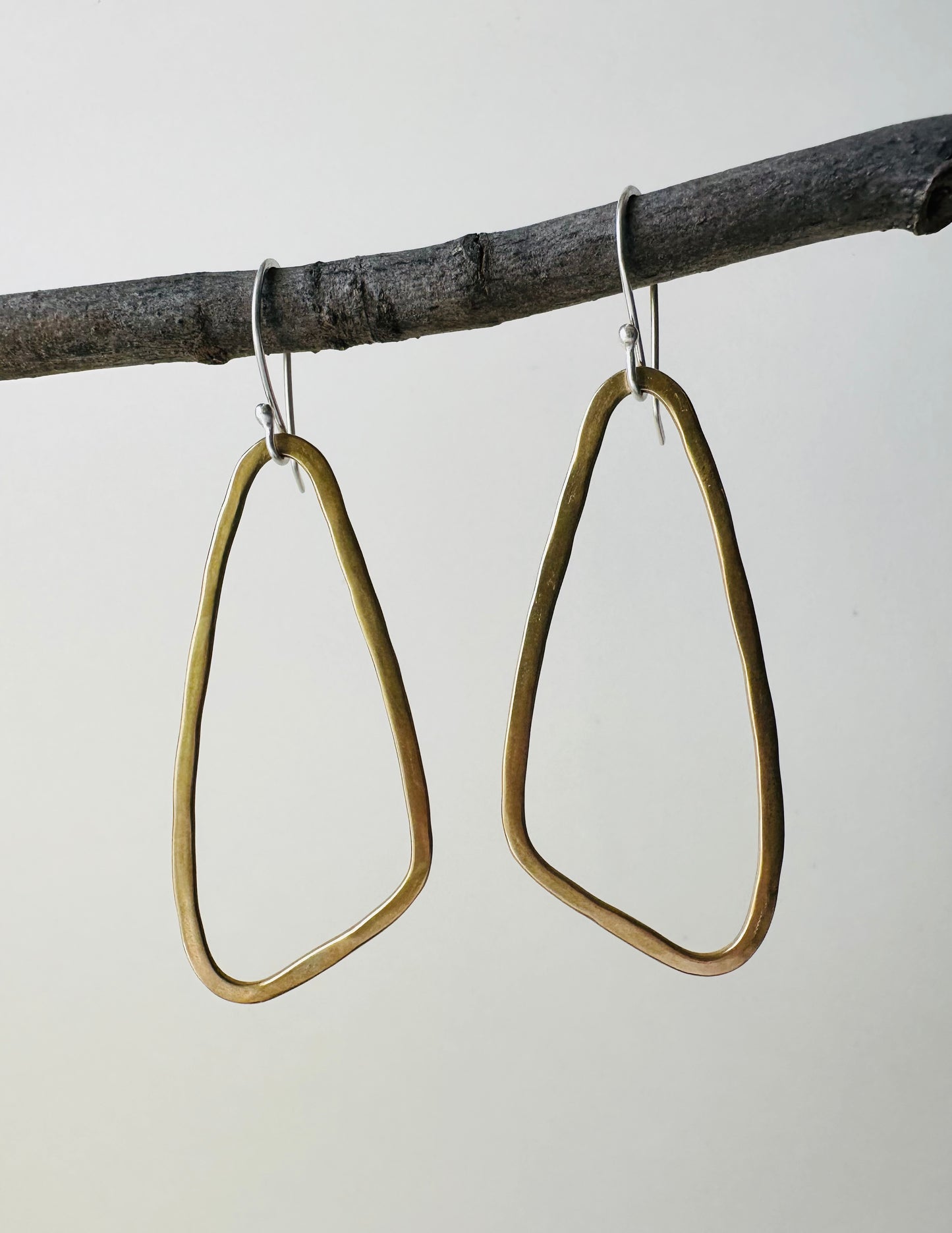 Brass wing shaped hoop earrings hanging on a branch against a neutral background