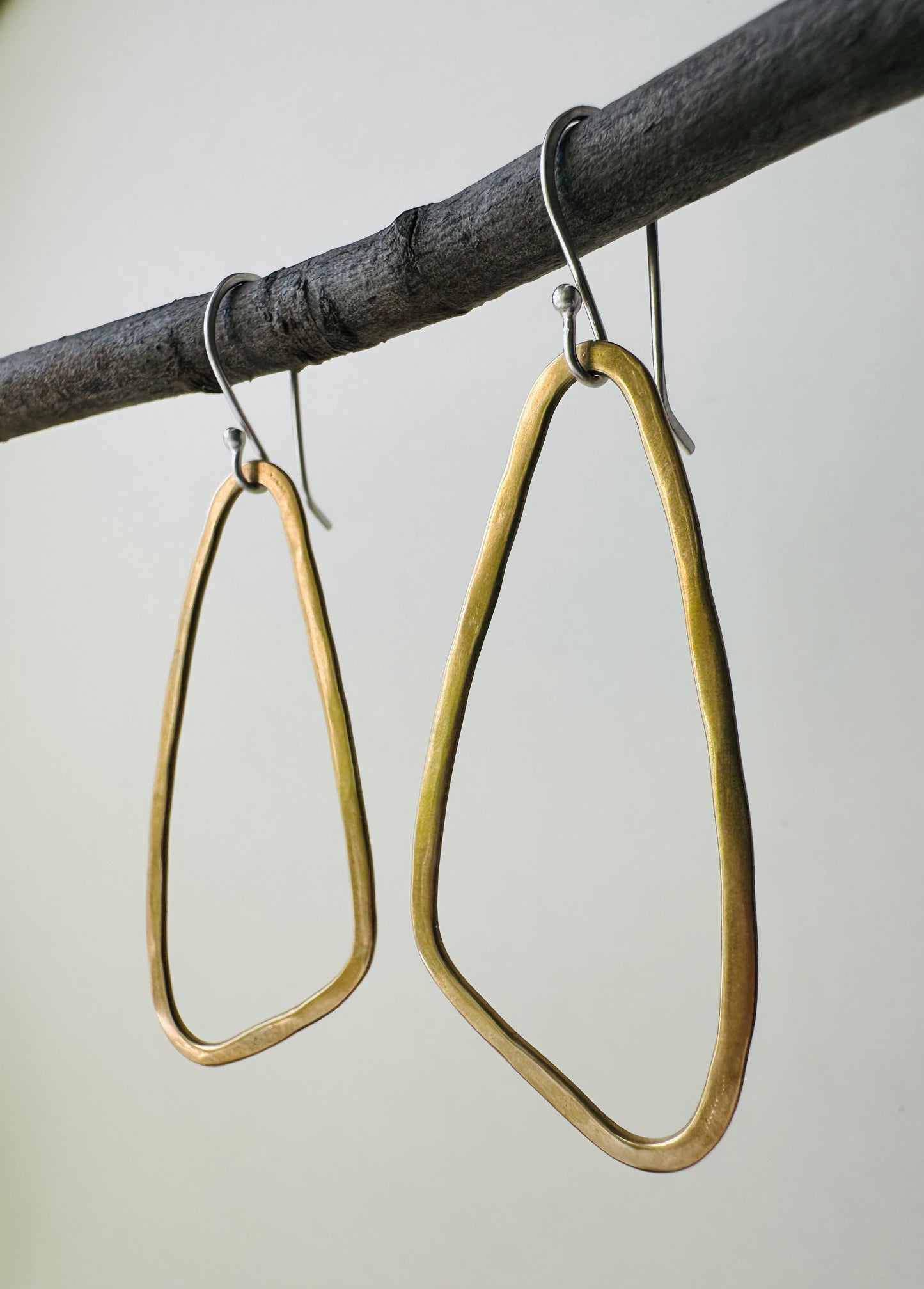 Brass wing shaped hoop earrings hanging on a branch against a neutral background