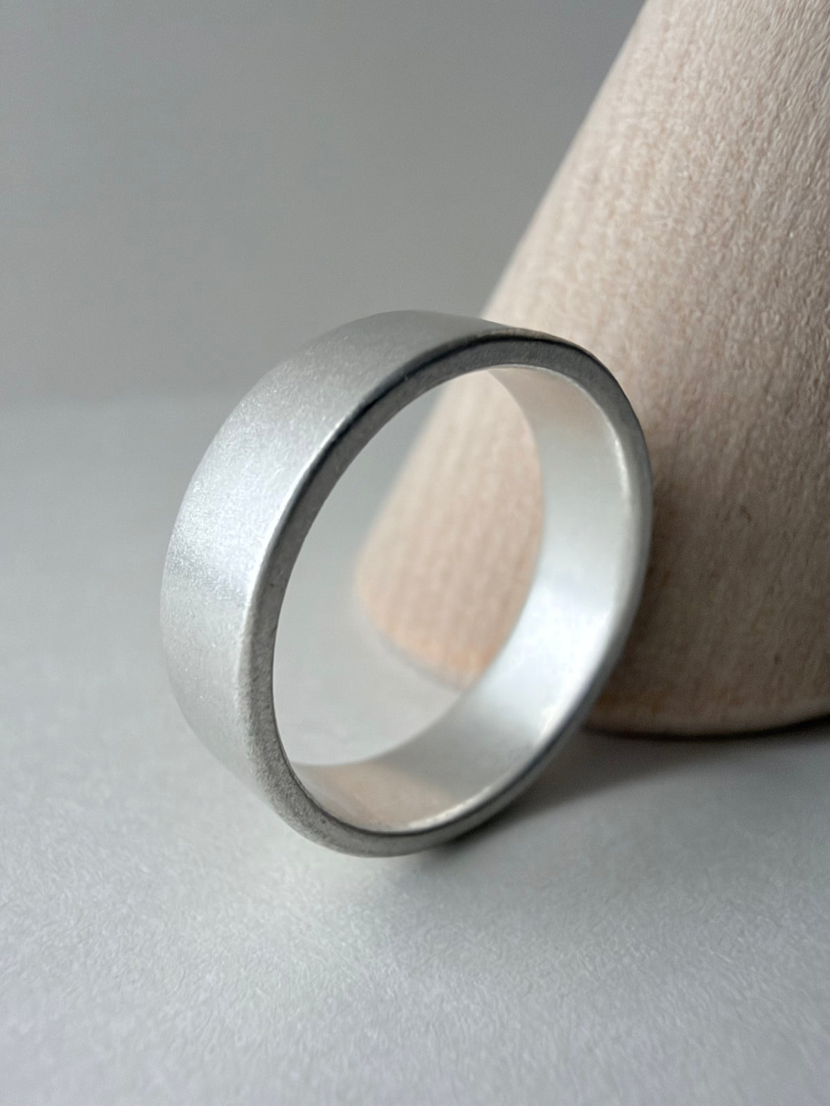 A minimalist silver band ring with a matte textured finish, resting on a light gray surface next to a wood cone display stand.
