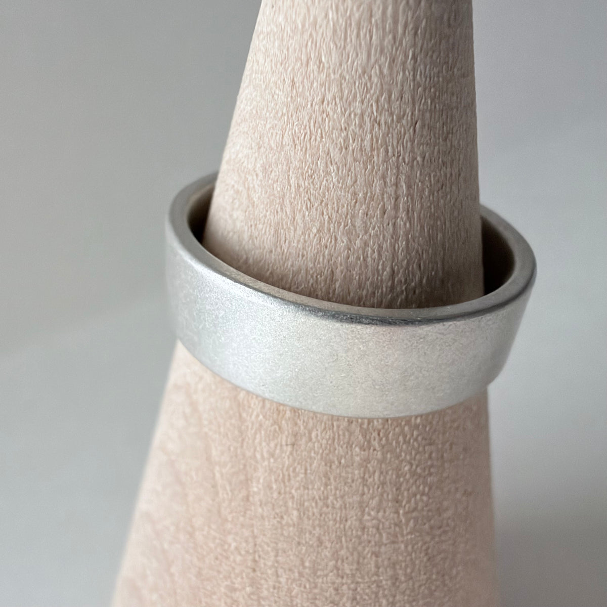 A wide, matte silver band ring displayed on a wood cone stand against a soft light gray background.
