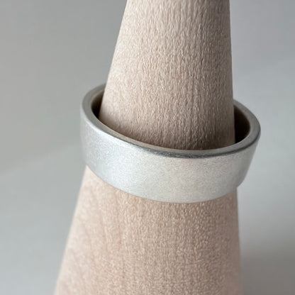 A wide, matte silver band ring displayed on a wood cone stand against a soft light gray background.