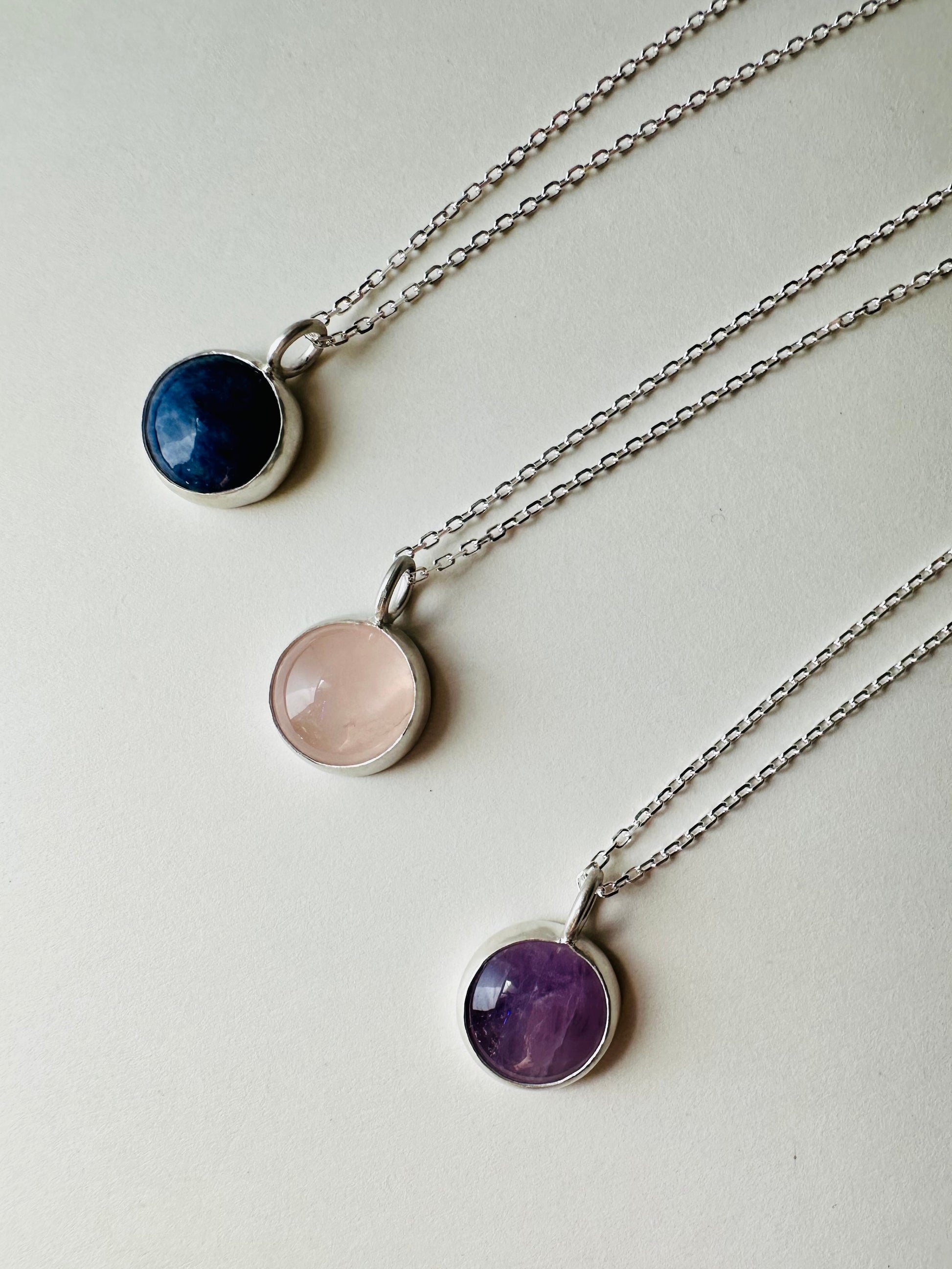 Three silver chain necklaces with round bezel-set gemstone pendants in navy blue, pale pink, and purple, arranged diagonally on a light off-white background.