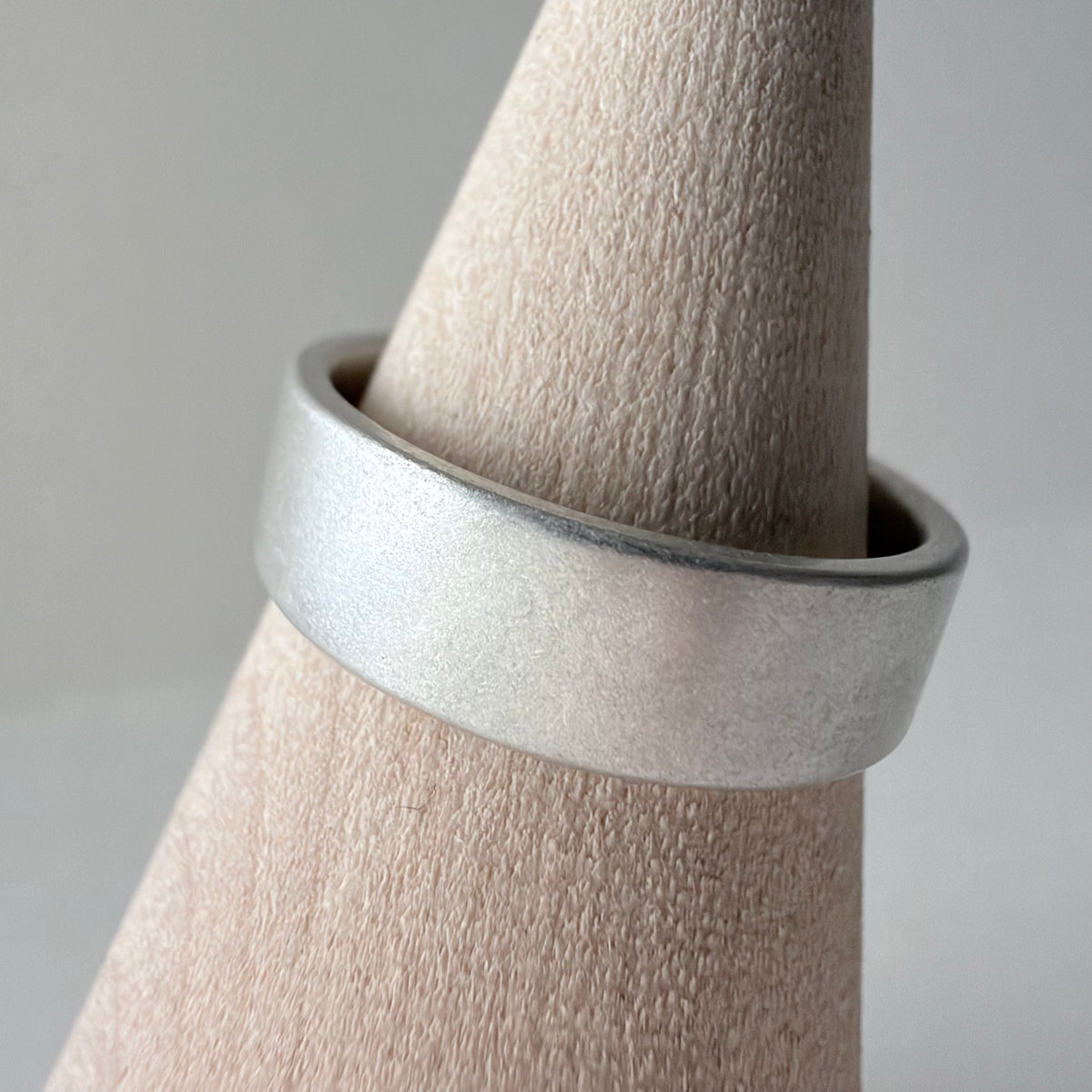 A wide, matte silver band ring displayed on a wood cone stand against a soft light gray background.