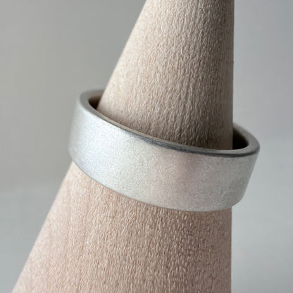 A wide, matte silver band ring displayed on a wood cone stand against a soft light gray background.