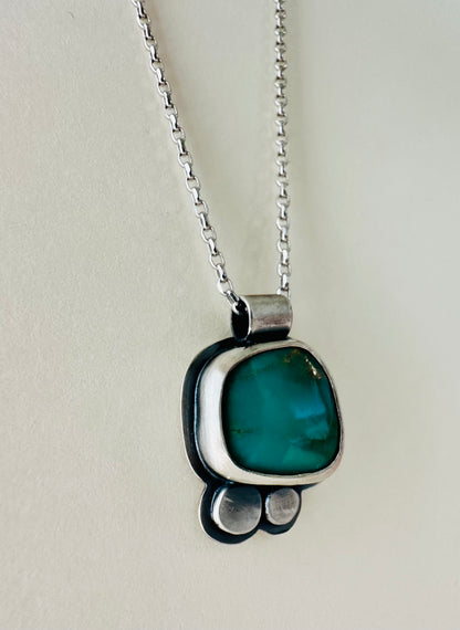 A sterling silver pendant necklace featuring a rounded organic shaped green stone, set in a silver bezel with two small silver dots below, hanging from a silver chain against a light cream background.