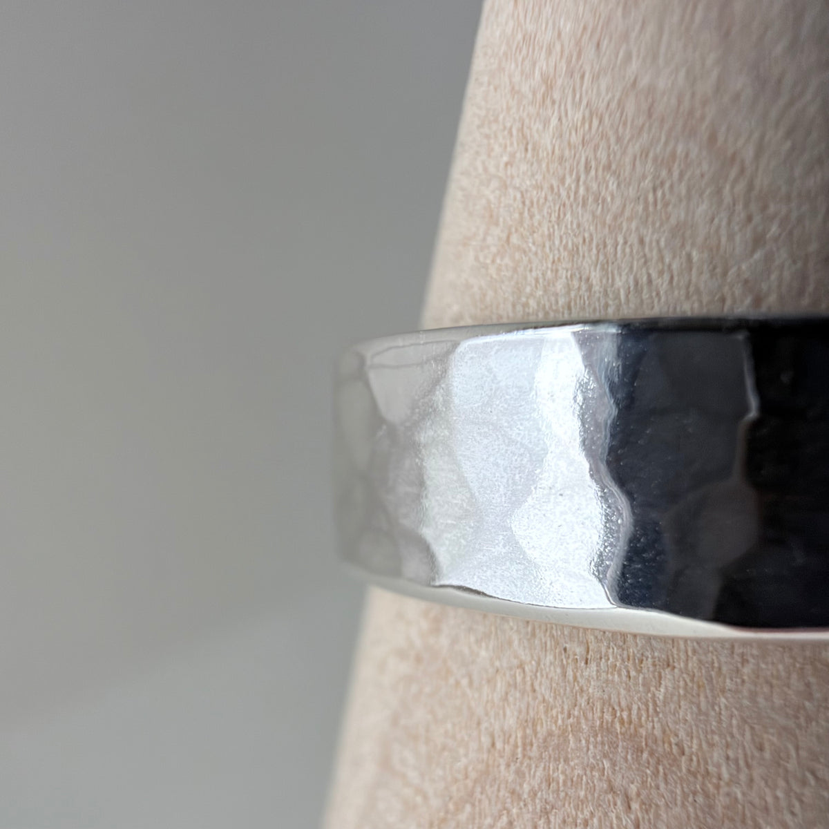 A close-up of a hammered silver band ring displayed on a wooden cone stand against a soft gray background.