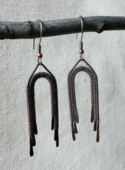 Close up image of copper wire wrapped rainbow dangle earrings on small branch. 